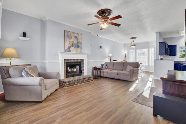 living room featuring a brick fireplace, hardwood / wood-style flooring, ceiling fan, ornamental molding, and a textured ceiling