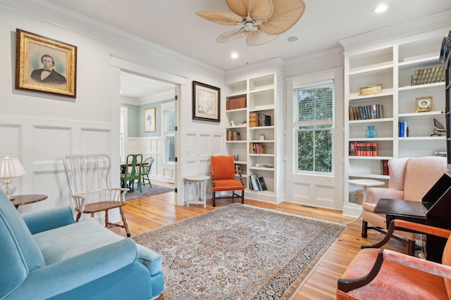 sitting room with a decorative wall, crown molding, wainscoting, light wood-style flooring, and ceiling fan