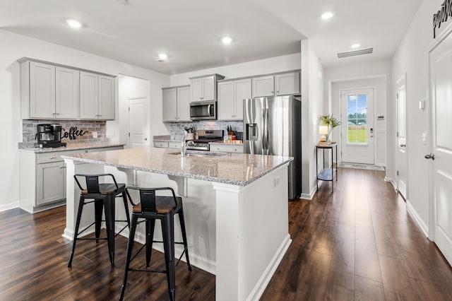 kitchen featuring appliances with stainless steel finishes, gray cabinetry, light stone counters, a center island with sink, and dark hardwood / wood-style floors