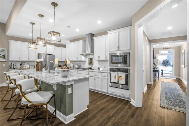 kitchen with appliances with stainless steel finishes, wall chimney range hood, decorative backsplash, a center island with sink, and white cabinetry