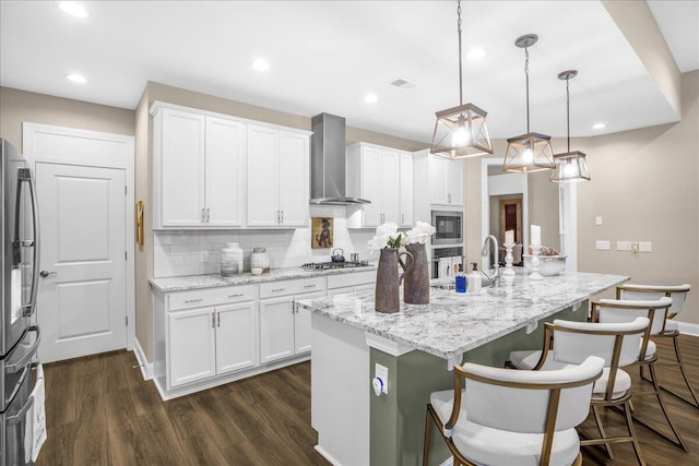 kitchen featuring wall chimney exhaust hood, a sink, appliances with stainless steel finishes, a kitchen bar, and white cabinets