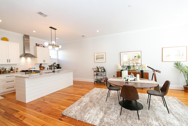 kitchen with white cabinetry, hanging light fixtures, a kitchen island with sink, light hardwood / wood-style floors, and wall chimney exhaust hood