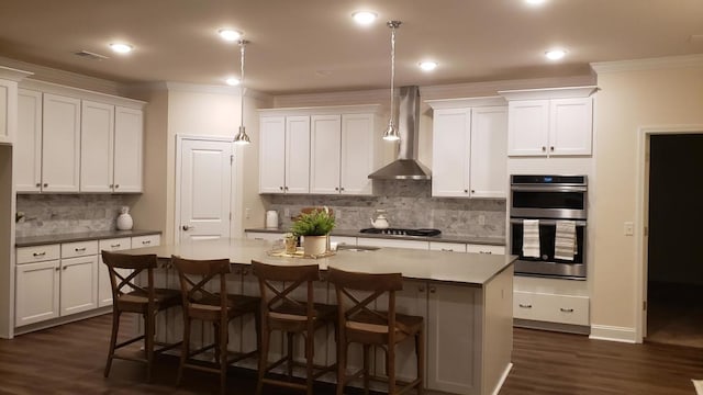 kitchen featuring white cabinets, double oven, a kitchen island with sink, and wall chimney exhaust hood