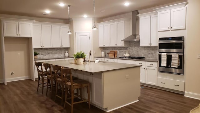 kitchen with a kitchen island with sink, white cabinets, wall chimney exhaust hood, decorative light fixtures, and stainless steel appliances