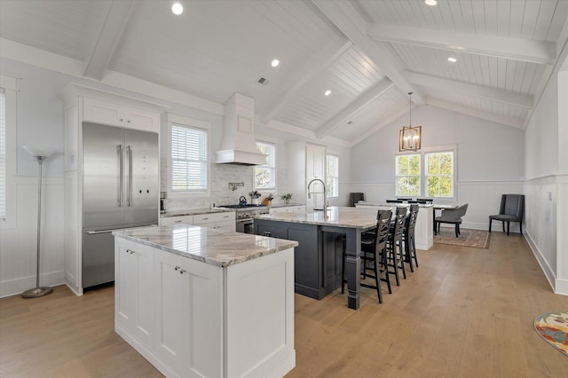 kitchen with custom exhaust hood, high end appliances, a center island with sink, light wood-type flooring, and white cabinetry