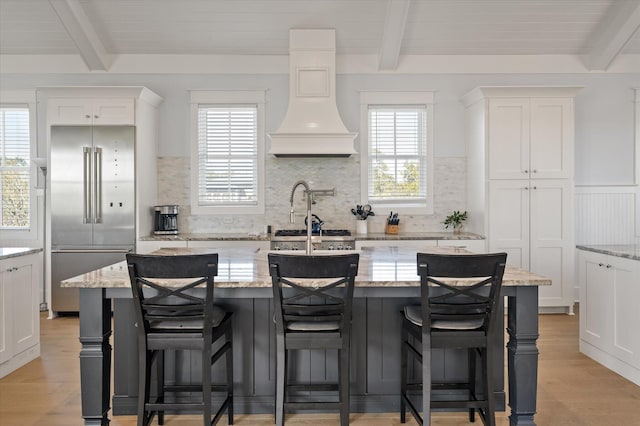 kitchen featuring beam ceiling, stainless steel appliances, light stone counters, and a kitchen island with sink