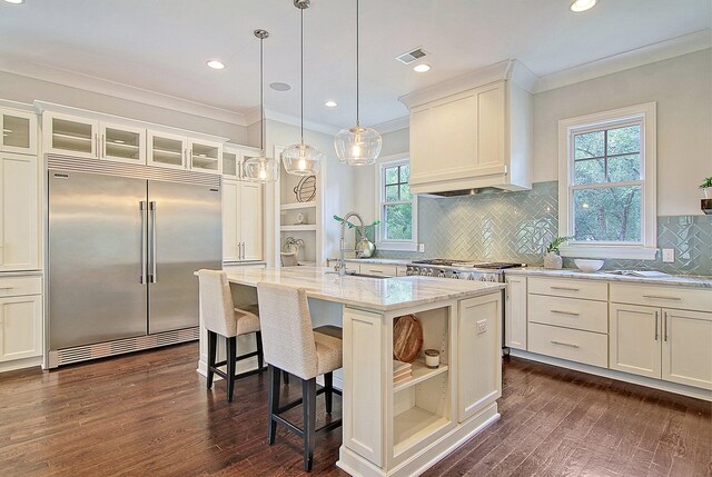 kitchen with built in refrigerator, dark wood-type flooring, pendant lighting, a center island with sink, and ornamental molding