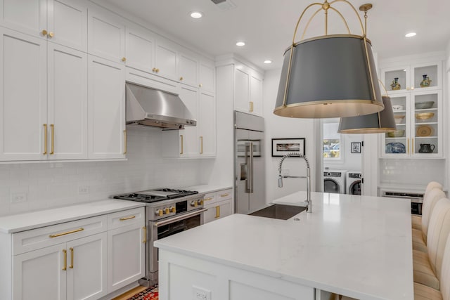 kitchen featuring wall chimney range hood, white cabinets, hanging light fixtures, washing machine and clothes dryer, and high quality appliances