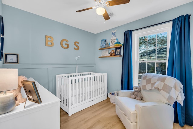 bedroom with ceiling fan, a decorative wall, a wainscoted wall, light wood-type flooring, and a crib