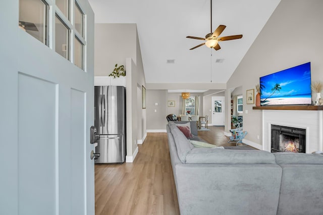 living room featuring a warm lit fireplace, baseboards, ceiling fan, light wood-type flooring, and high vaulted ceiling