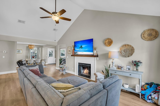 living area featuring a warm lit fireplace, visible vents, light wood-style flooring, ceiling fan, and high vaulted ceiling