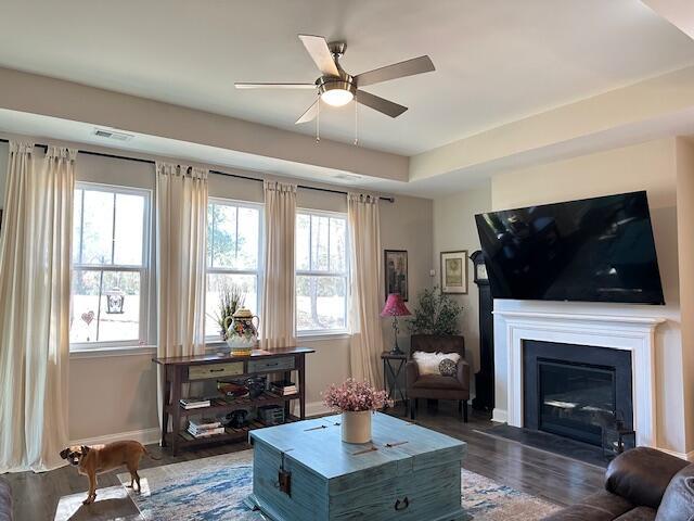 living room featuring a tray ceiling, dark wood-style flooring, a glass covered fireplace, ceiling fan, and baseboards