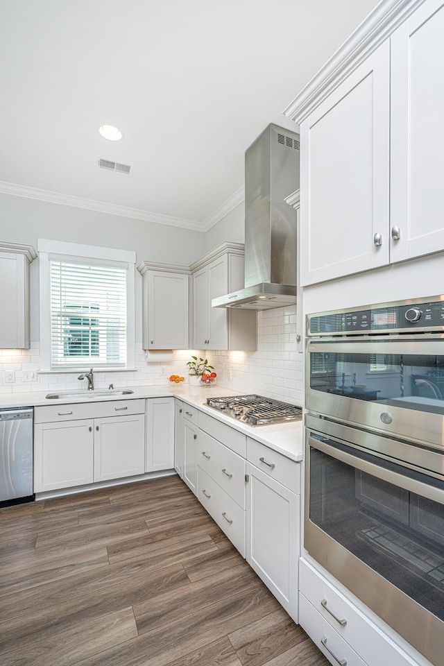 kitchen with wall chimney range hood, a sink, stainless steel appliances, ornamental molding, and visible vents