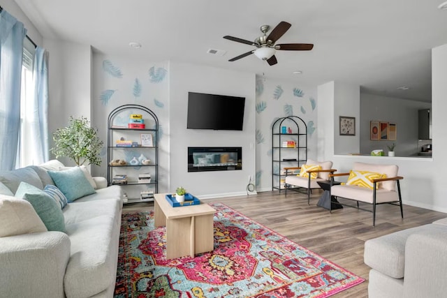 living room with ceiling fan, wood finished floors, a glass covered fireplace, and baseboards