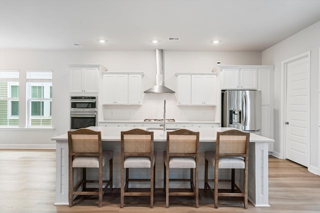 kitchen with wall chimney exhaust hood, stainless steel appliances, light countertops, an island with sink, and white cabinetry