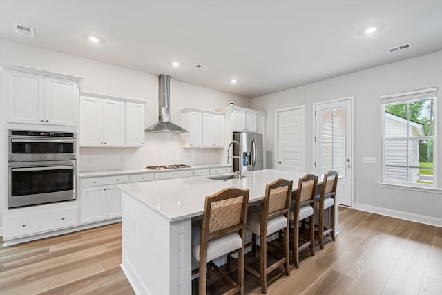 kitchen featuring wall chimney range hood, stainless steel appliances, a sink, an island with sink, and light wood finished floors