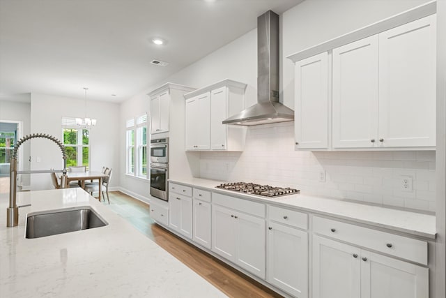 kitchen with wall chimney range hood, a sink, white cabinetry, a chandelier, and wood finished floors