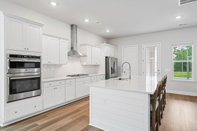 kitchen with stainless steel appliances, wall chimney range hood, a sink, a kitchen island with sink, and light wood-type flooring