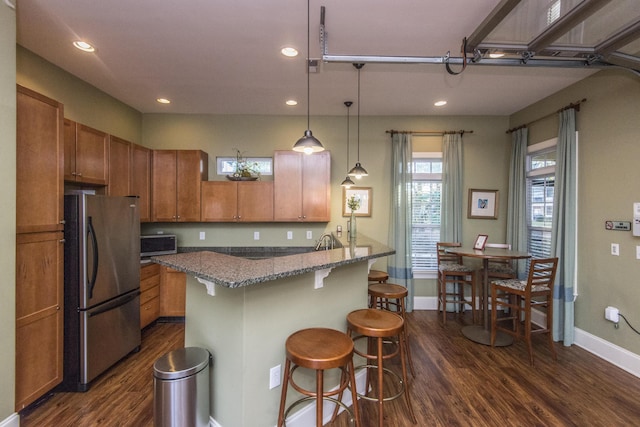 kitchen with dark wood finished floors, light stone countertops, brown cabinetry, appliances with stainless steel finishes, and a breakfast bar area