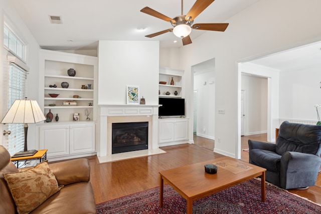 living room with built in shelves, ceiling fan, wood-type flooring, and vaulted ceiling