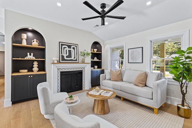 living room featuring a tile fireplace, vaulted ceiling, light hardwood / wood-style flooring, ceiling fan, and built in shelves