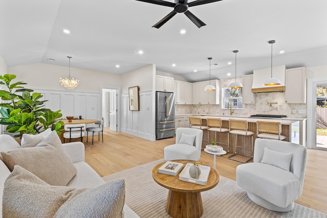 living room with sink, ceiling fan with notable chandelier, light hardwood / wood-style floors, and vaulted ceiling