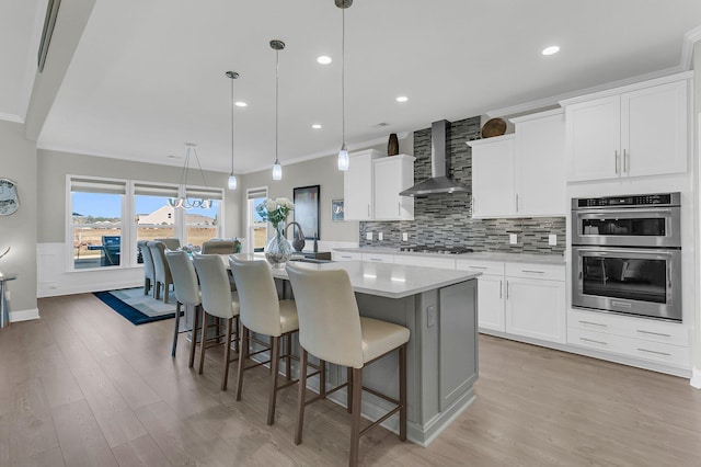 kitchen featuring white cabinetry, wall chimney range hood, an island with sink, and pendant lighting