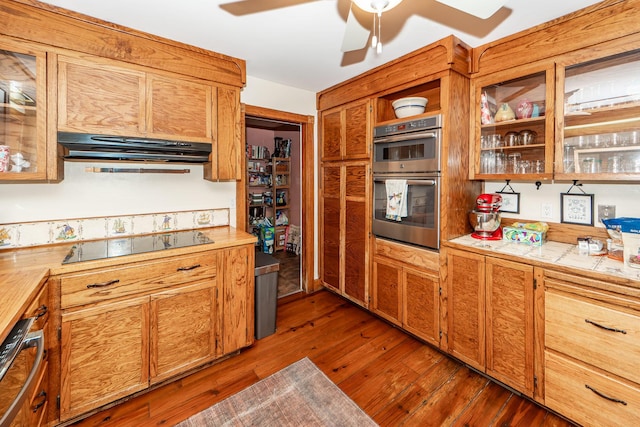 kitchen with dark wood-style floors, black electric stovetop, light countertops, stainless steel double oven, and under cabinet range hood