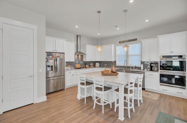 kitchen featuring stainless steel appliances, wall chimney range hood, pendant lighting, white cabinets, and light hardwood / wood-style floors