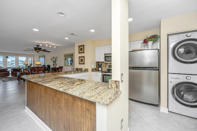 kitchen featuring stacked washer and dryer, white cabinetry, stainless steel appliances, light stone counters, and kitchen peninsula