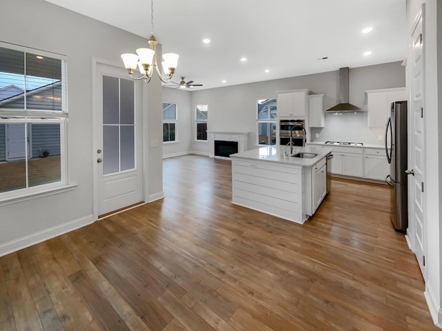 kitchen with white cabinets, wall chimney range hood, an island with sink, decorative light fixtures, and stovetop