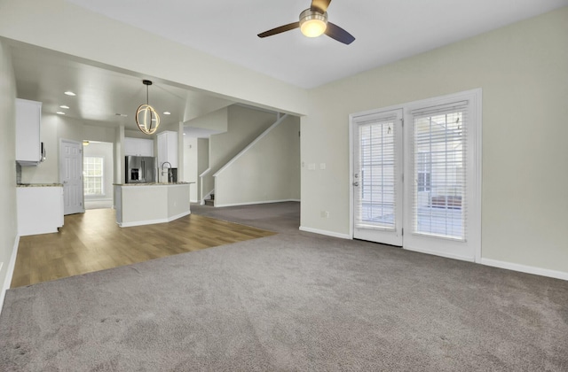 unfurnished living room with ceiling fan, a sink, baseboards, stairway, and dark colored carpet