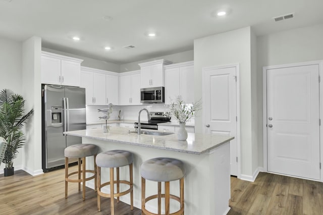 kitchen with hardwood / wood-style floors, stainless steel appliances, light stone countertops, a kitchen island with sink, and white cabinets