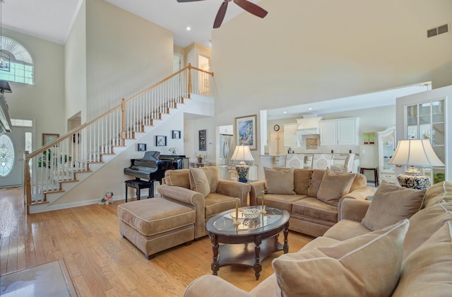living area featuring a towering ceiling, stairs, light wood-style flooring, ceiling fan, and baseboards