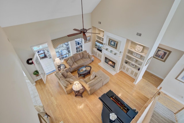 living area with high vaulted ceiling, a ceiling fan, built in shelves, wood finished floors, and a lit fireplace
