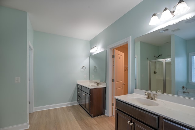 bathroom featuring wood finished floors, a stall shower, two vanities, baseboards, and a walk in closet