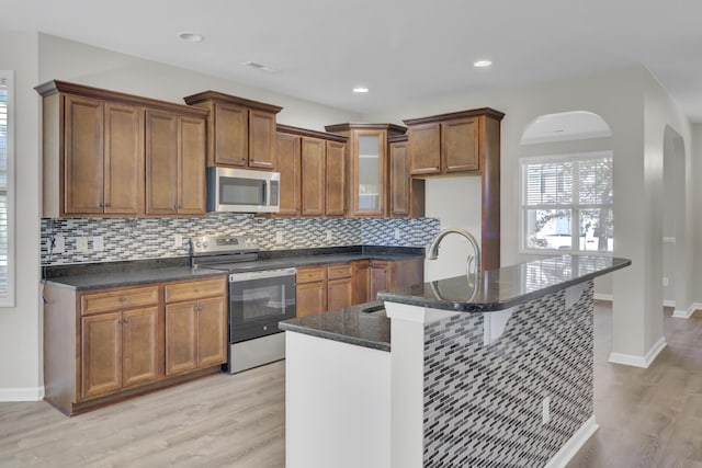 kitchen featuring dark stone counters, a center island with sink, light hardwood / wood-style flooring, appliances with stainless steel finishes, and tasteful backsplash