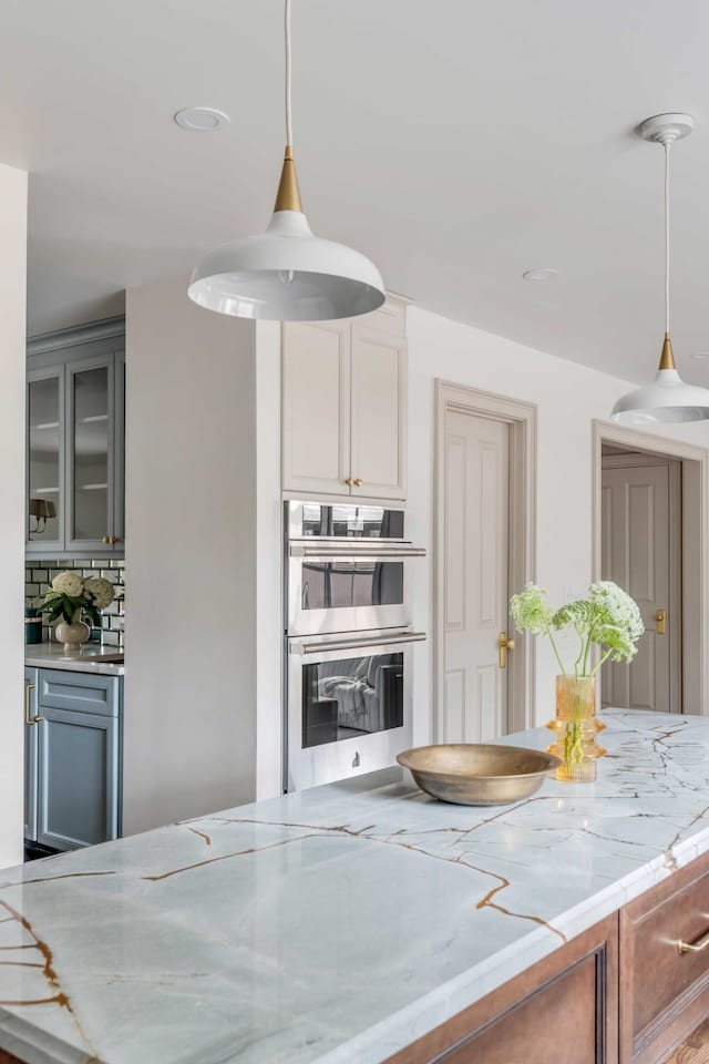 kitchen featuring light stone countertops, double oven, backsplash, decorative light fixtures, and gray cabinets