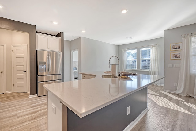 kitchen with stainless steel fridge, white cabinets, an island with sink, light wood-style flooring, and a sink