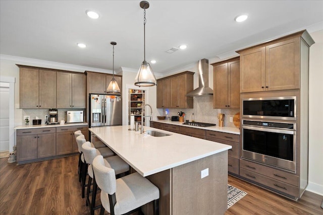 kitchen with wall chimney range hood, dark wood-type flooring, a center island with sink, sink, and stainless steel appliances