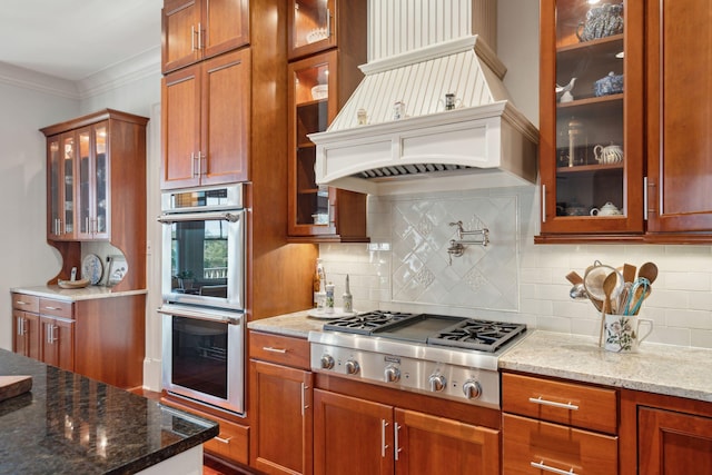 kitchen with glass insert cabinets, appliances with stainless steel finishes, light stone counters, custom exhaust hood, and crown molding