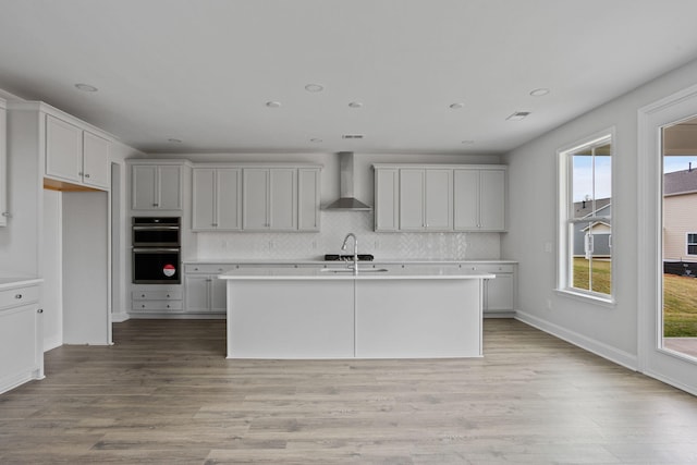 kitchen featuring an island with sink, light wood-style flooring, double oven, wall chimney exhaust hood, and decorative backsplash