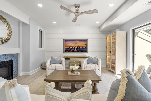 living room with ceiling fan, a tiled fireplace, light hardwood / wood-style flooring, and wood walls