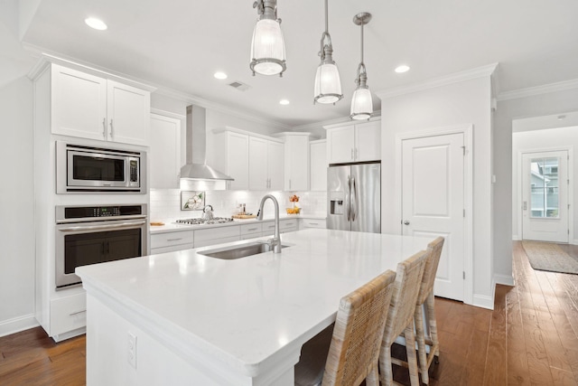 kitchen with appliances with stainless steel finishes, sink, hanging light fixtures, a kitchen island with sink, and wall chimney exhaust hood