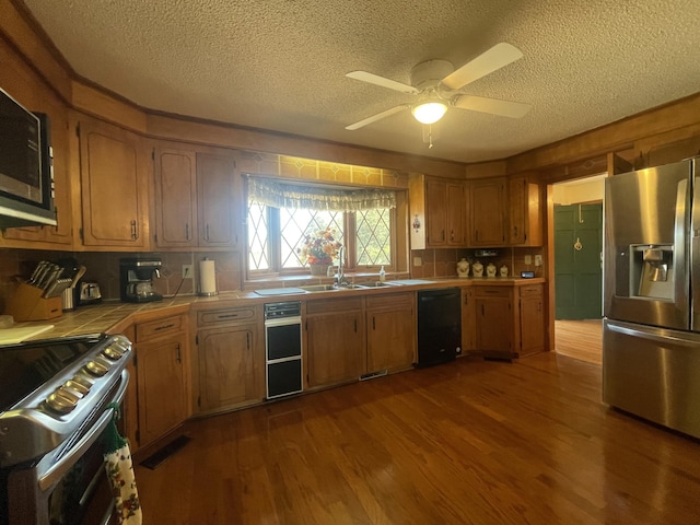 kitchen featuring brown cabinetry, tile countertops, appliances with stainless steel finishes, dark wood-style flooring, and a sink