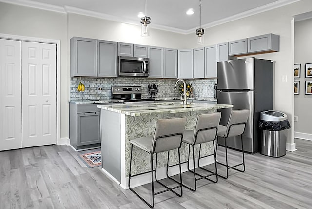 kitchen featuring gray cabinetry, a kitchen island with sink, light stone counters, stainless steel appliances, and crown molding