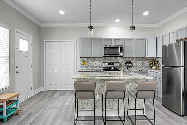 kitchen featuring crown molding, appliances with stainless steel finishes, a kitchen island with sink, hanging light fixtures, and light stone countertops