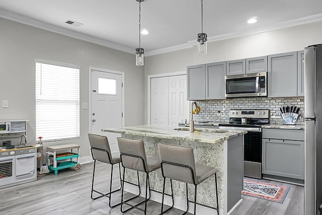 kitchen with a kitchen island with sink, hanging light fixtures, gray cabinetry, stainless steel appliances, and light stone countertops