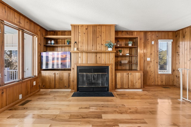 unfurnished living room with wooden walls, light hardwood / wood-style flooring, built in shelves, ornamental molding, and a textured ceiling