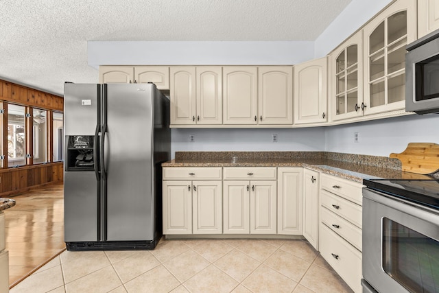 kitchen with a textured ceiling, stainless steel appliances, light tile patterned floors, dark stone countertops, and cream cabinetry